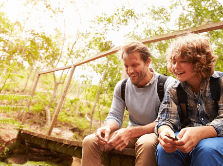 A man and a boy sitting on a wooden bridge in a forested area. The man, wearing a light blue sweater, and the boy, dressed in a plaid shirt, are smiling while holding small objects. Lush greenery surrounds them, creating a natural setting.