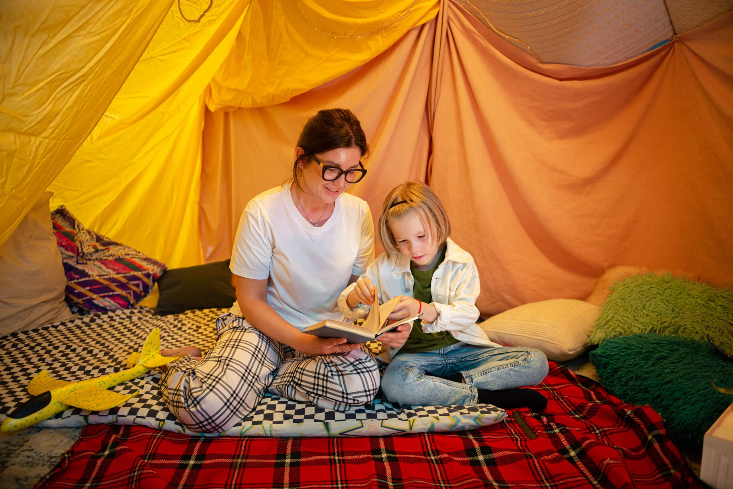A woman in glasses and a child sitting together in a cozy indoor tent, reading a book. The tent has yellow and pink fabric draping, and the ground is covered with checkered and plaid blankets. The atmosphere appears warm and inviting.