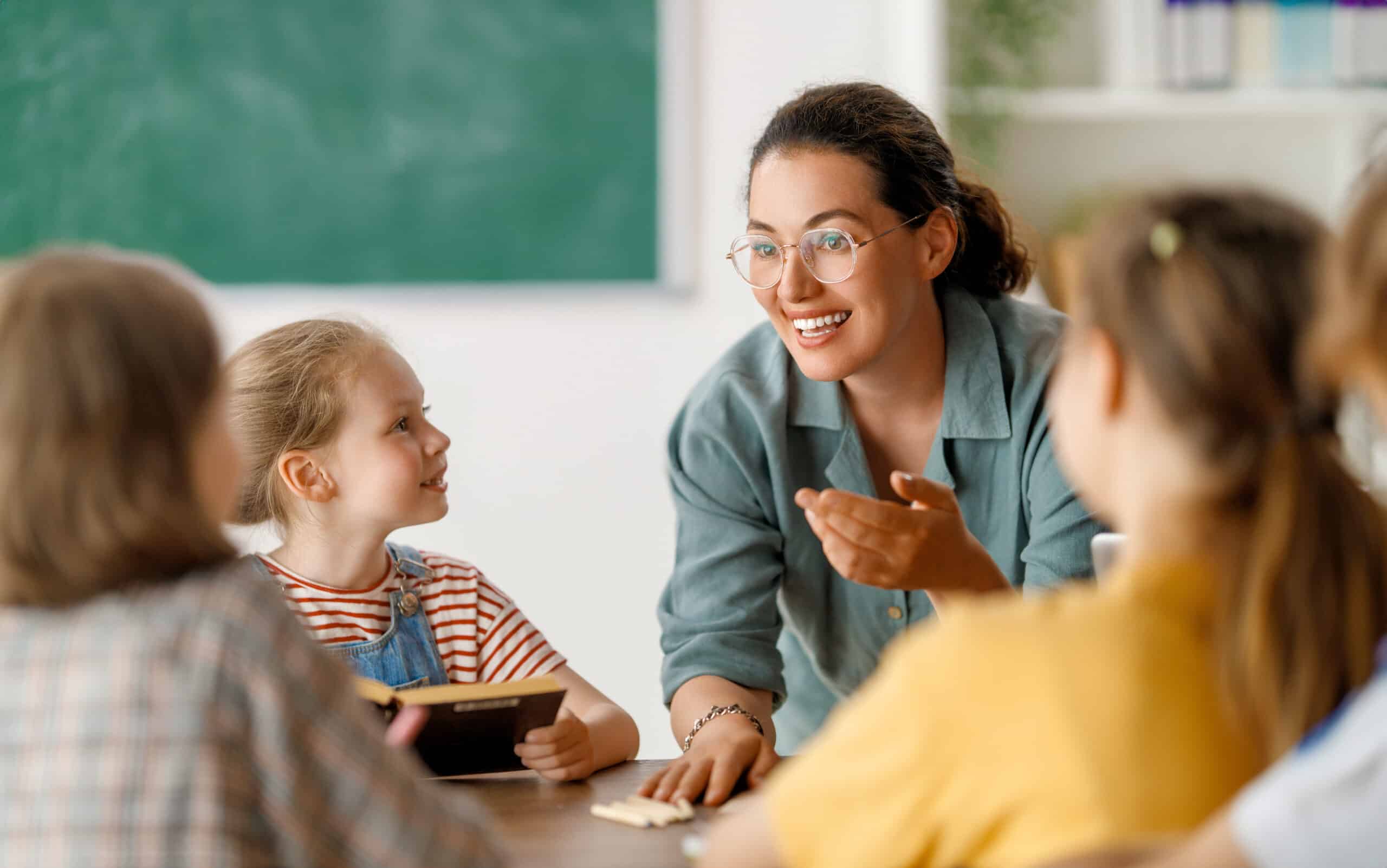 A teacher interacts with a group of children at a classroom table. The teacher, wearing glasses, is smiling and gesturing while speaking. The children, including one girl in a striped shirt, listen attentively, showing a collaborative learning environment.