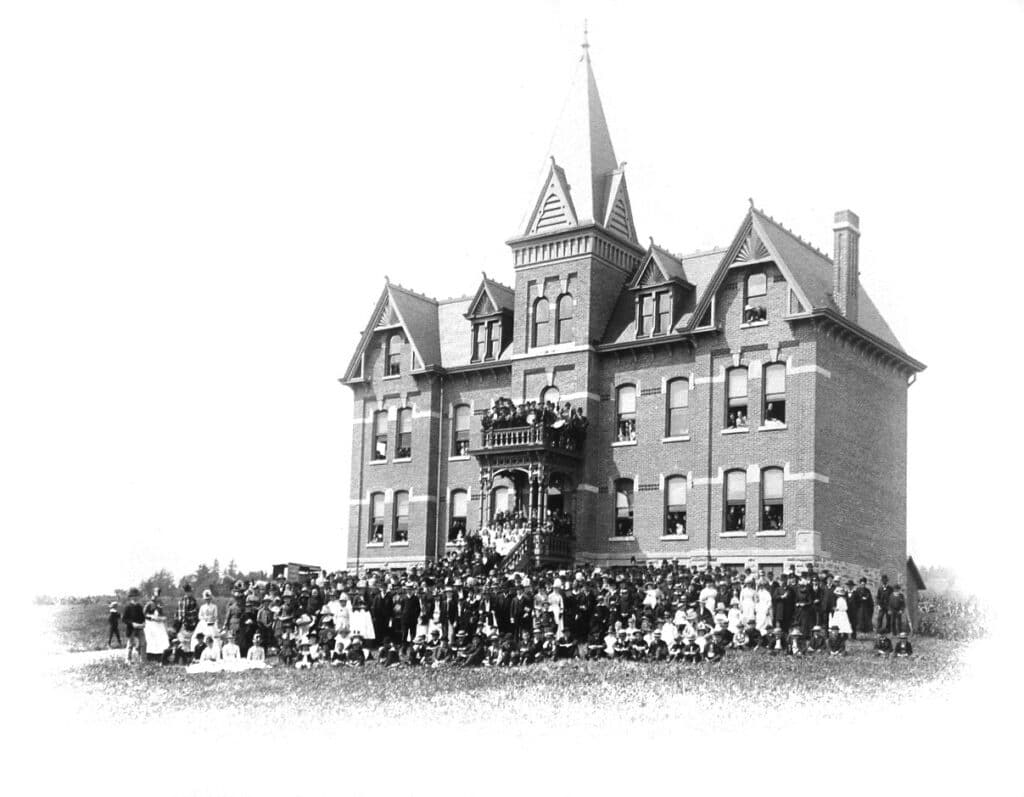 A historic brick mansion with a tall spire, surrounded by a large group of people, dressed in early 20th-century clothing. The individuals stand on the lawn in front of the building, which features multiple windows and decorative architecture.