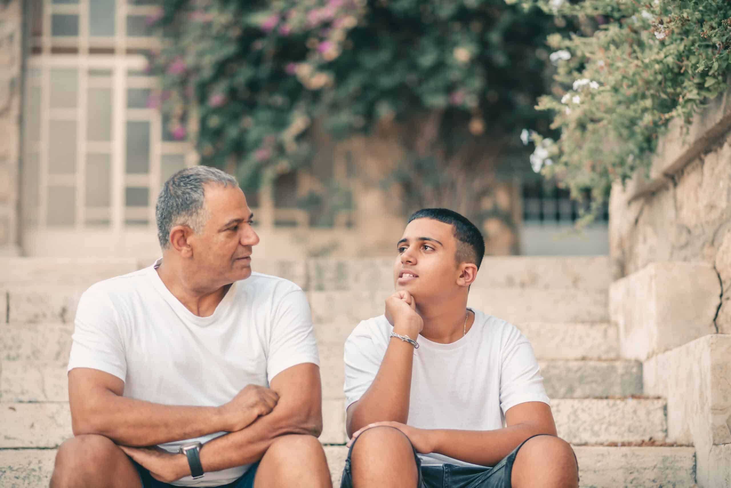 A middle-aged man and a teenage boy sit on stone steps in a relaxed atmosphere. They are both wearing casual white t-shirts. The man looks attentively at the boy, who is engaged in thought and looking away, surrounded by greenery and flowers.