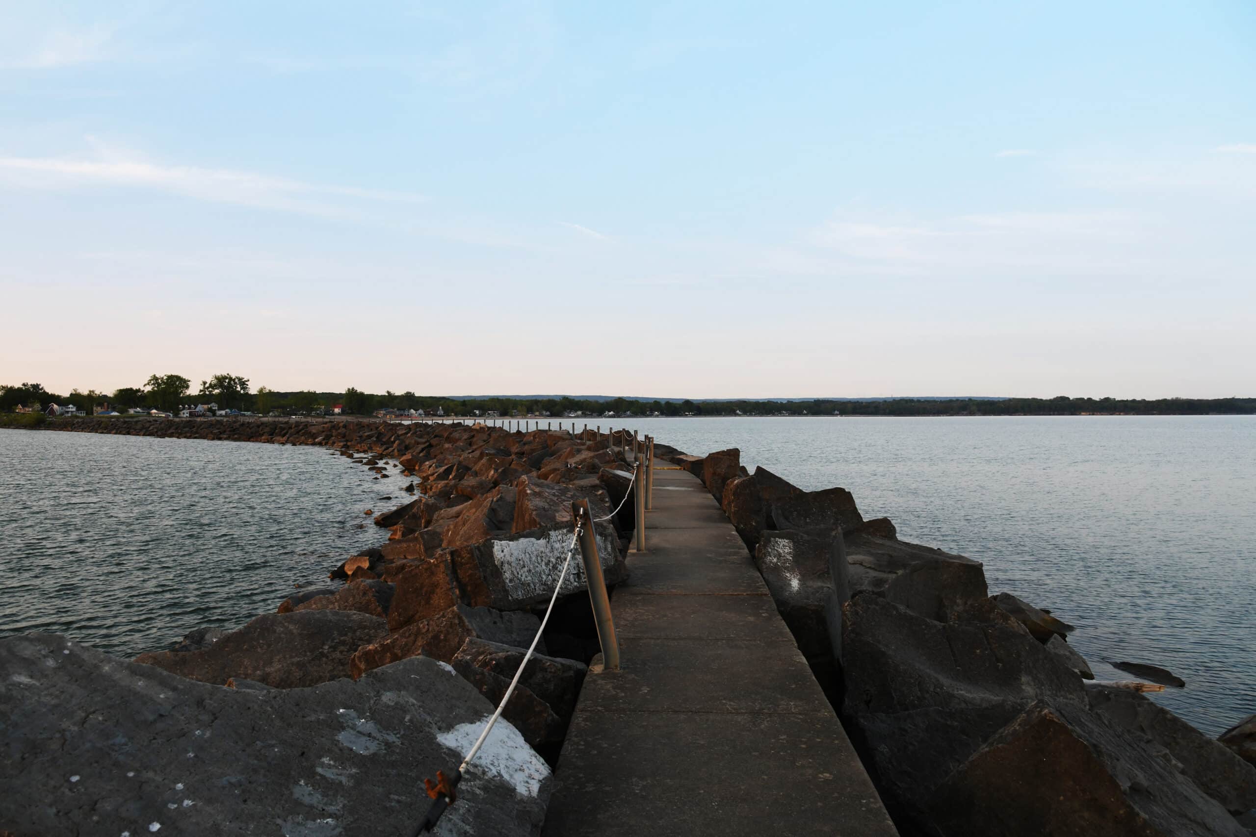 A concrete path leads out over a rocky jetty into calm water, with the shoreline visible in the distance. The sky is clear with a setting sun, creating a soft blue and orange hue. Small rocks line the edges of the path, and a railing is present on one side.