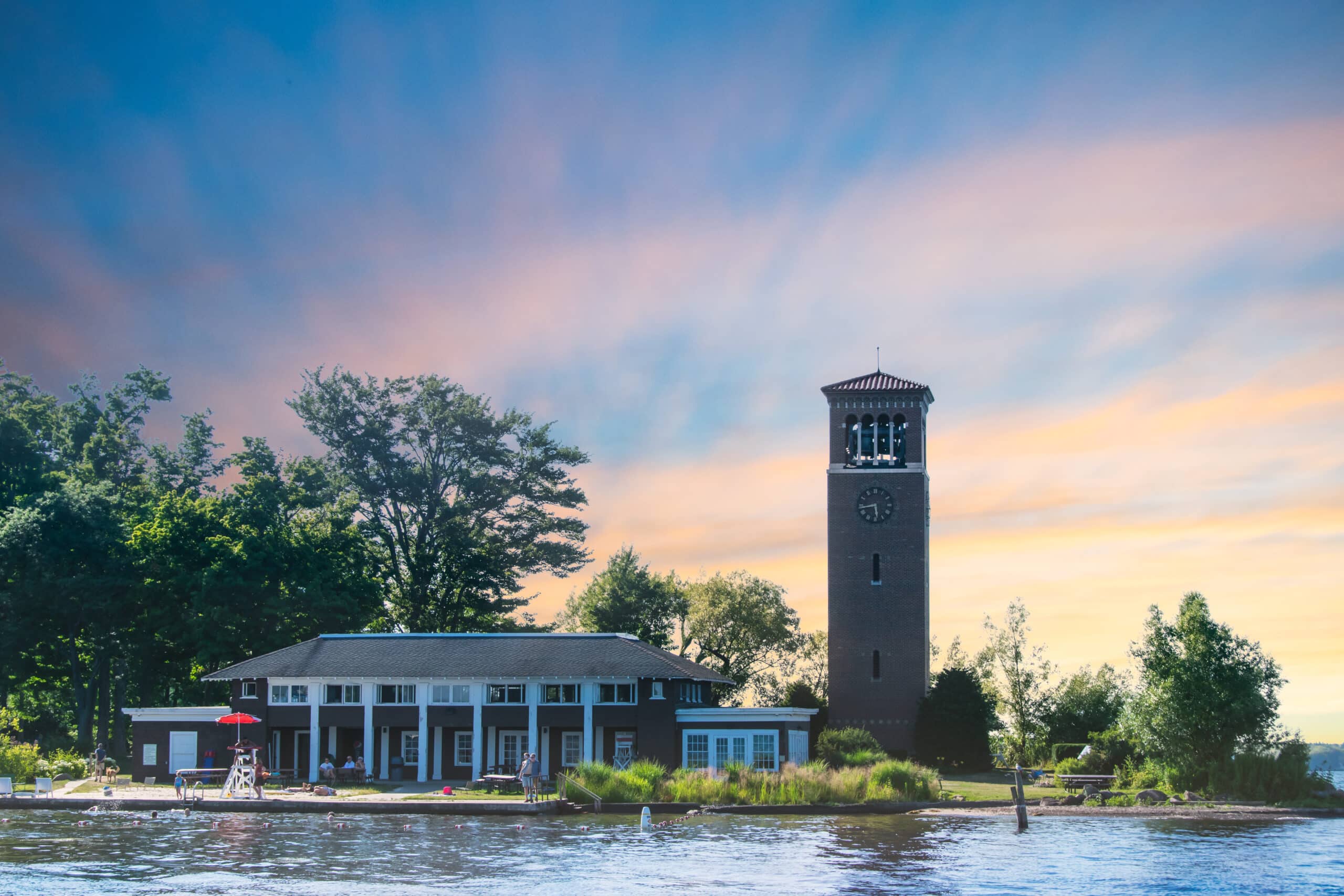 A lakeside building with a clock tower, surrounded by trees and green foliage. The building features large windows and an outdoor area with a lifeguard chair. The scene is set against a colorful sky during sunset.