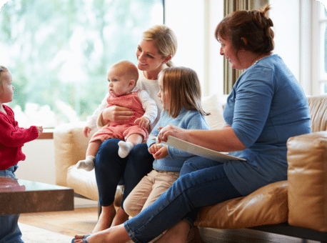 A woman holding a baby sits on a couch with two children beside her. A woman in blue is seated across from them, engaging with the children. The setting is a brightly lit living room with a large window in the background.