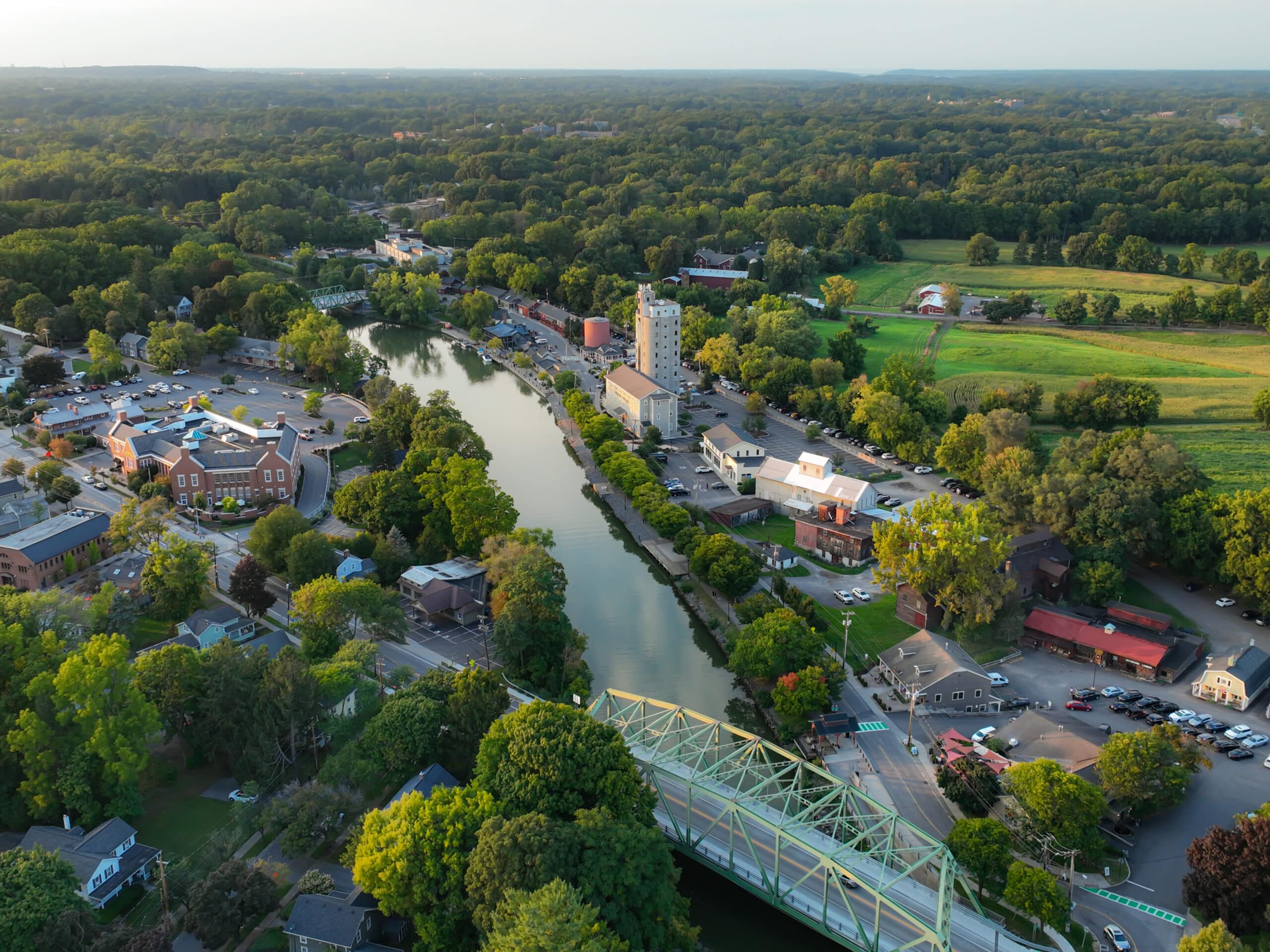 Aerial view of a small town featuring a river running through the center, surrounded by trees and greenery. There are several buildings along the riverbanks, including shops and homes, along with a bridge crossing the river.