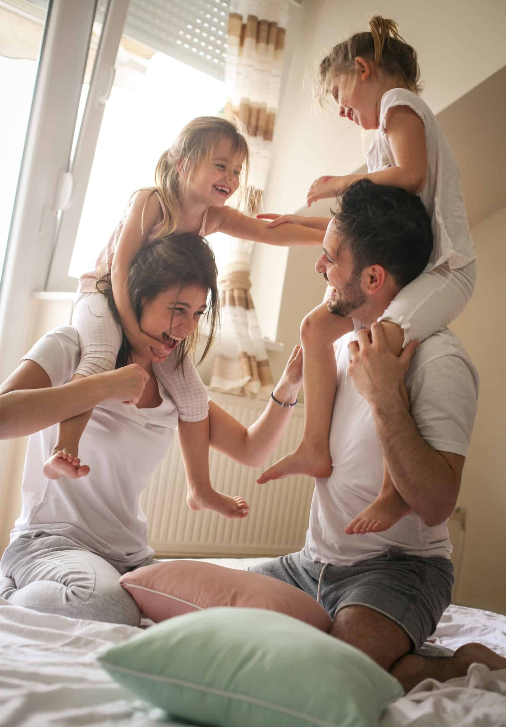 A family of four playing together in a brightly lit room. The father is giving a piggyback ride to one daughter, while the mother is playfully engaging with the other daughter beside her. Soft pillows are scattered on the bed, enhancing the playful atmosphere.