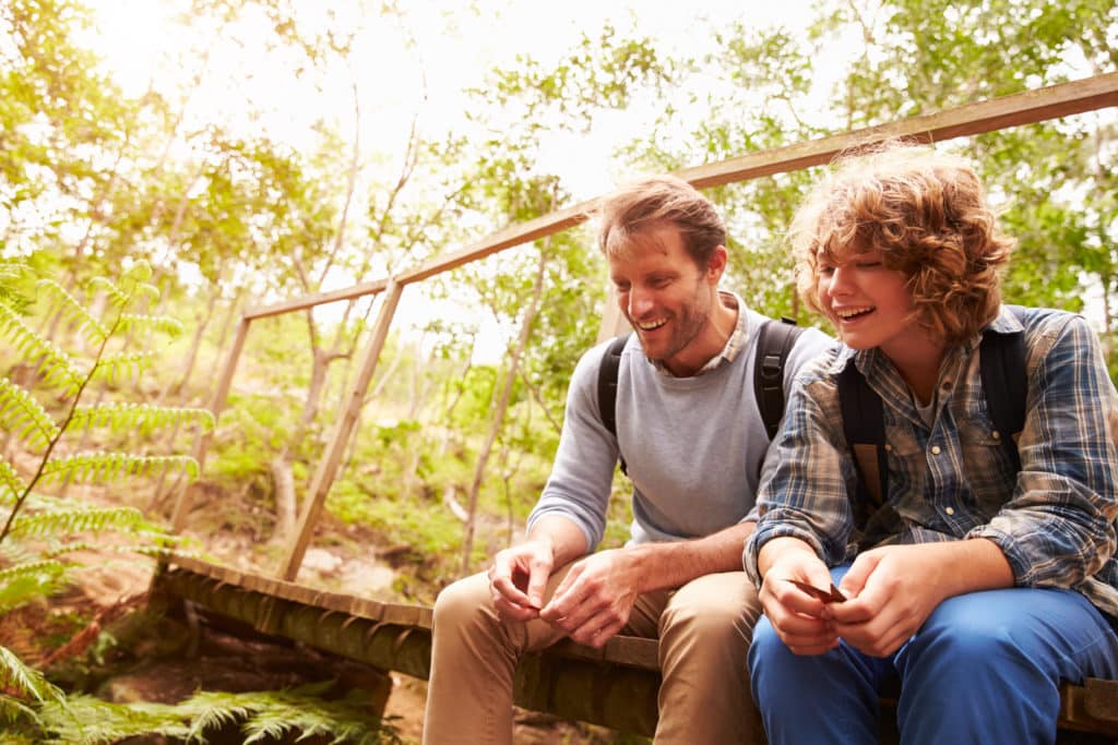 A man and a boy sitting on a wooden bridge in a forested area, surrounded by greenery. The man has short hair and is wearing a sweater, and the boy has curly hair and is dressed in a plaid shirt. Both are smiling and engaged in conversation.