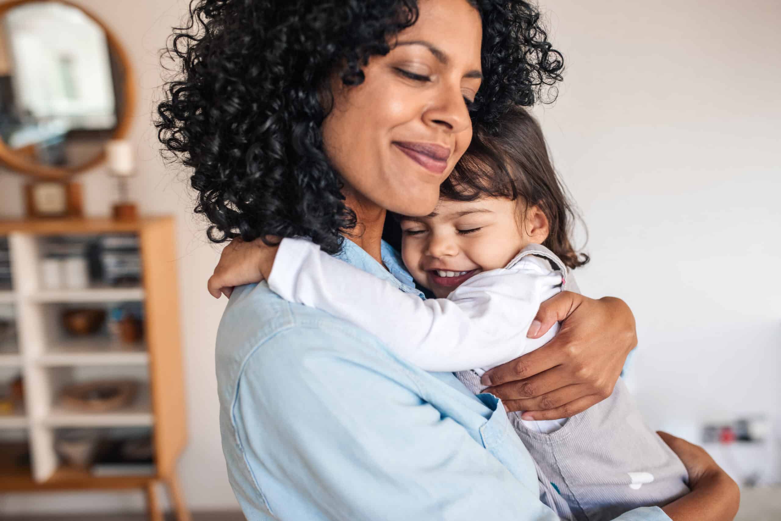 A woman with curly hair hugs a young girl with brown hair. They are both smiling and appear to share a close, affectionate moment in a bright, modern living space with wooden furniture.