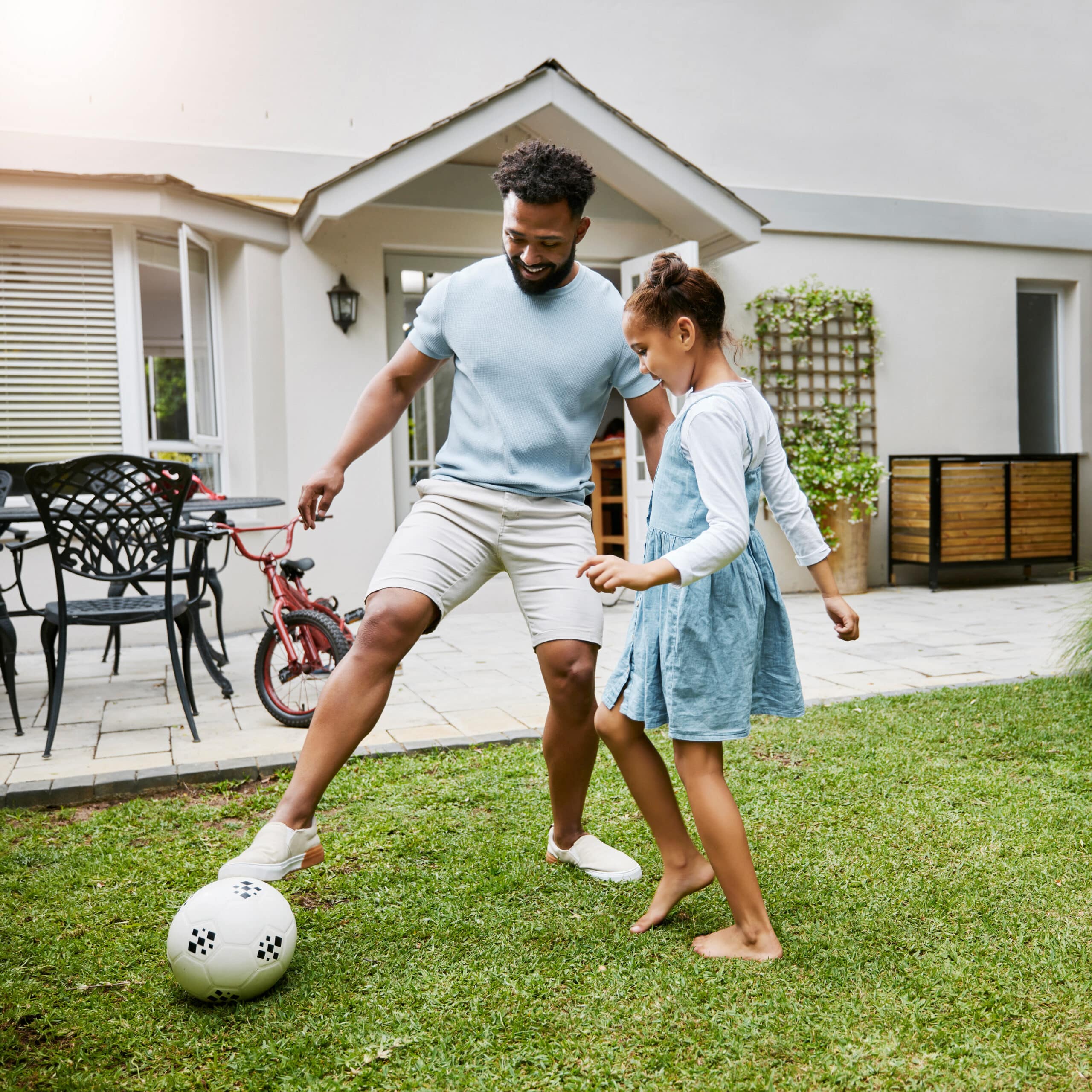 A man and a girl play soccer in a grassy backyard. The man, wearing a light blue shirt and shorts, playfully nudges a black and white soccer ball with one foot. The girl, in a blue dress with a white shirt underneath, watches attentively.
