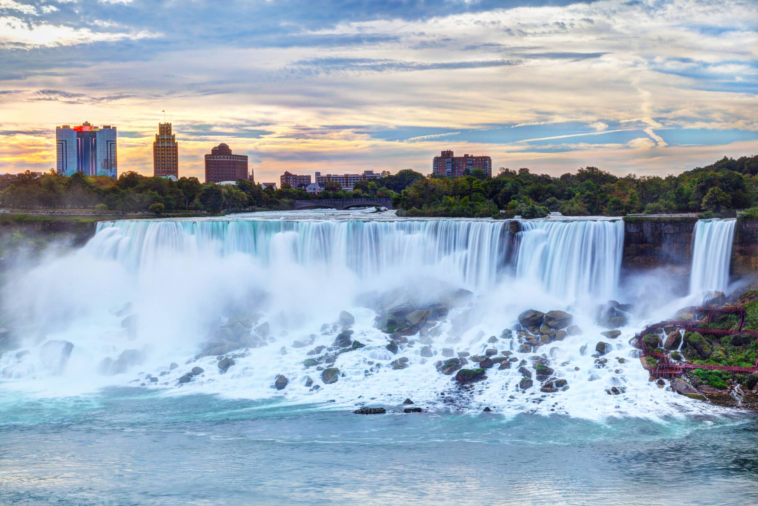 Niagara Falls with water cascading dramatically into the river below. Surrounding the falls are lush greenery and rocks. In the background, the skyline of a city can be seen under a partly cloudy sky near sunset.