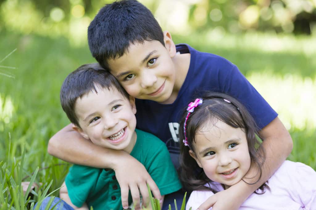 Three children sitting together on grass in a garden. A boy in a blue shirt is hugging two younger kids. One boy in a green shirt is smiling, while a girl in a pink shirt has her hair adorned with a bow. The background is blurred with greenery.