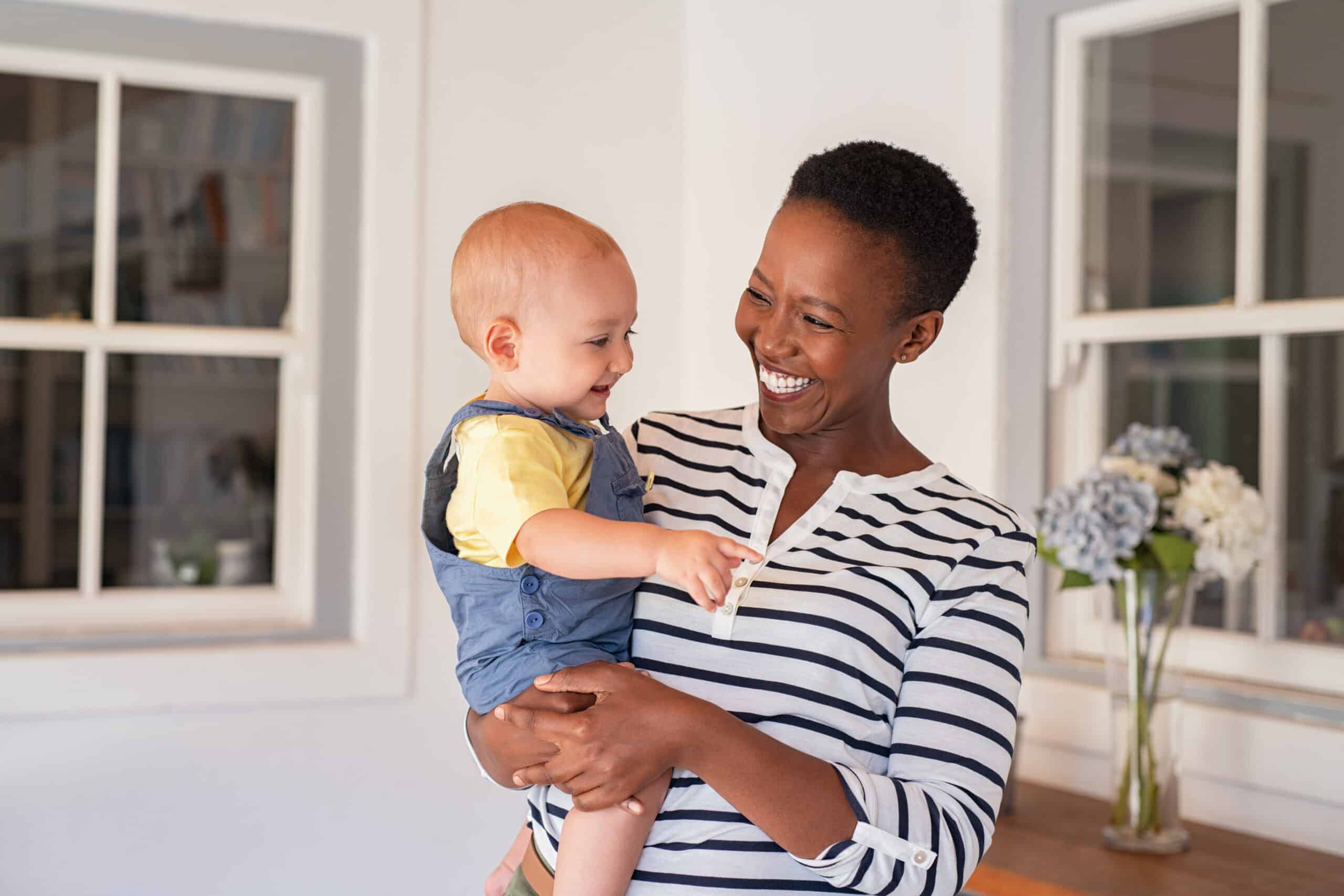 A woman with a short hairstyle is smiling while holding a baby in her arms. The baby, wearing a blue outfit with yellow accents, is looking at the woman. They are in a bright room with white walls and a wooden table, decorated with flowers.