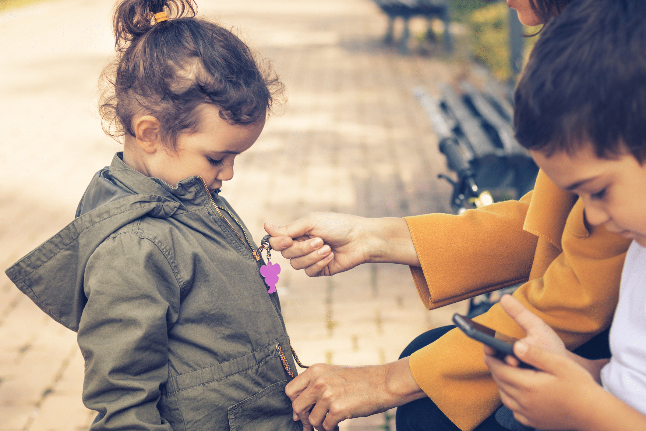 A woman assists a young girl in zipping her jacket while seated on a park bench. The girl has curly hair and is wearing a green jacket, and a boy nearby is focused on a smartphone.