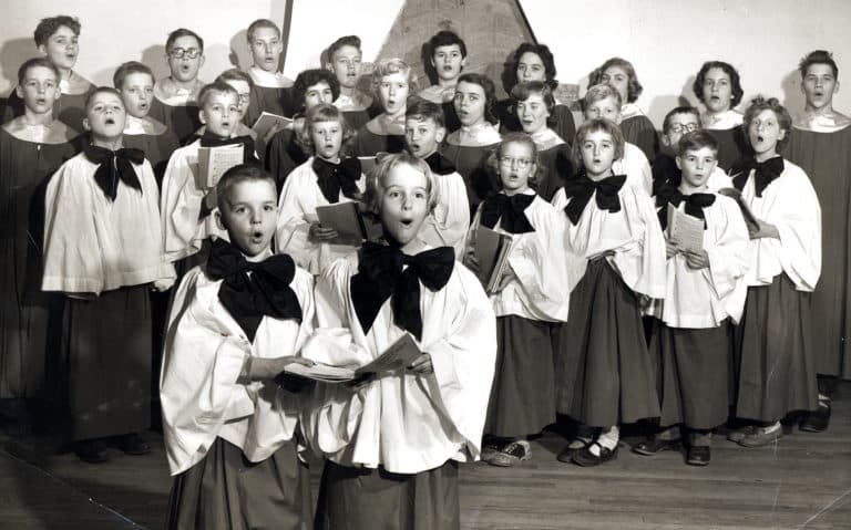 A black-and-white photo of a choir composed of children wearing matching choir robes with large bows at the neck. They are arranged in rows, some holding sheet music. The background features a simple stage setting.