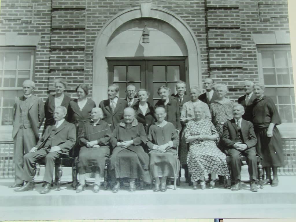 A black-and-white photo of a large group of men and women posing in front of a brick building. The group includes both seated and standing individuals, dressed in vintage clothing, with smiles and neutral expressions.