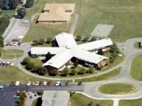 Aerial view of a school campus featuring a unique star-shaped building surrounded by green fields. Additional buildings and a parking lot are visible nearby, along with a circular driveway for vehicle access.