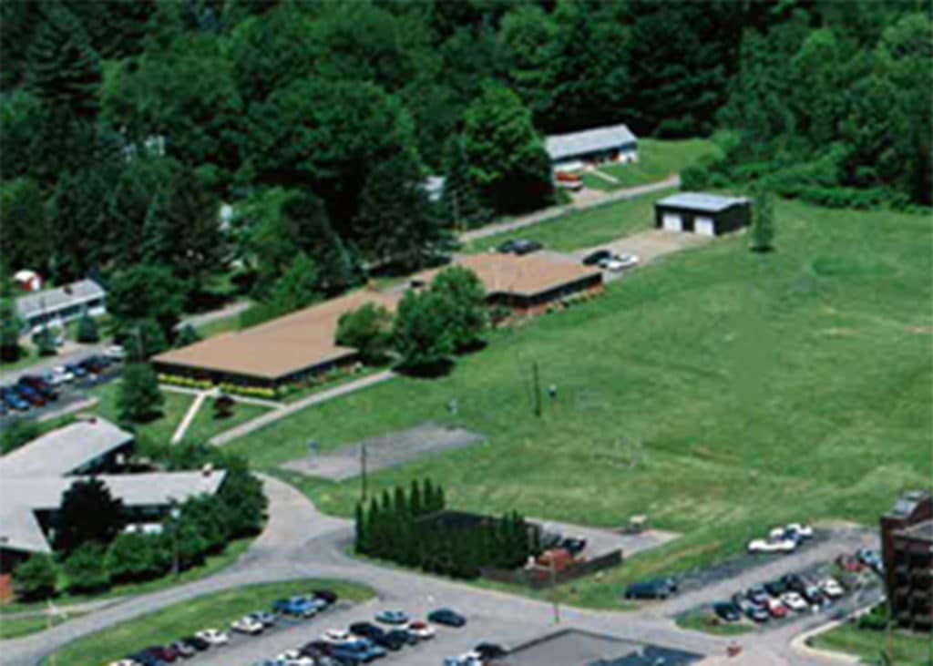 Aerial view of a landscaped area featuring several buildings, including a larger structure with a brown roof, surrounded by grassy fields and trees. Multiple parked cars are visible in a parking lot nearby.