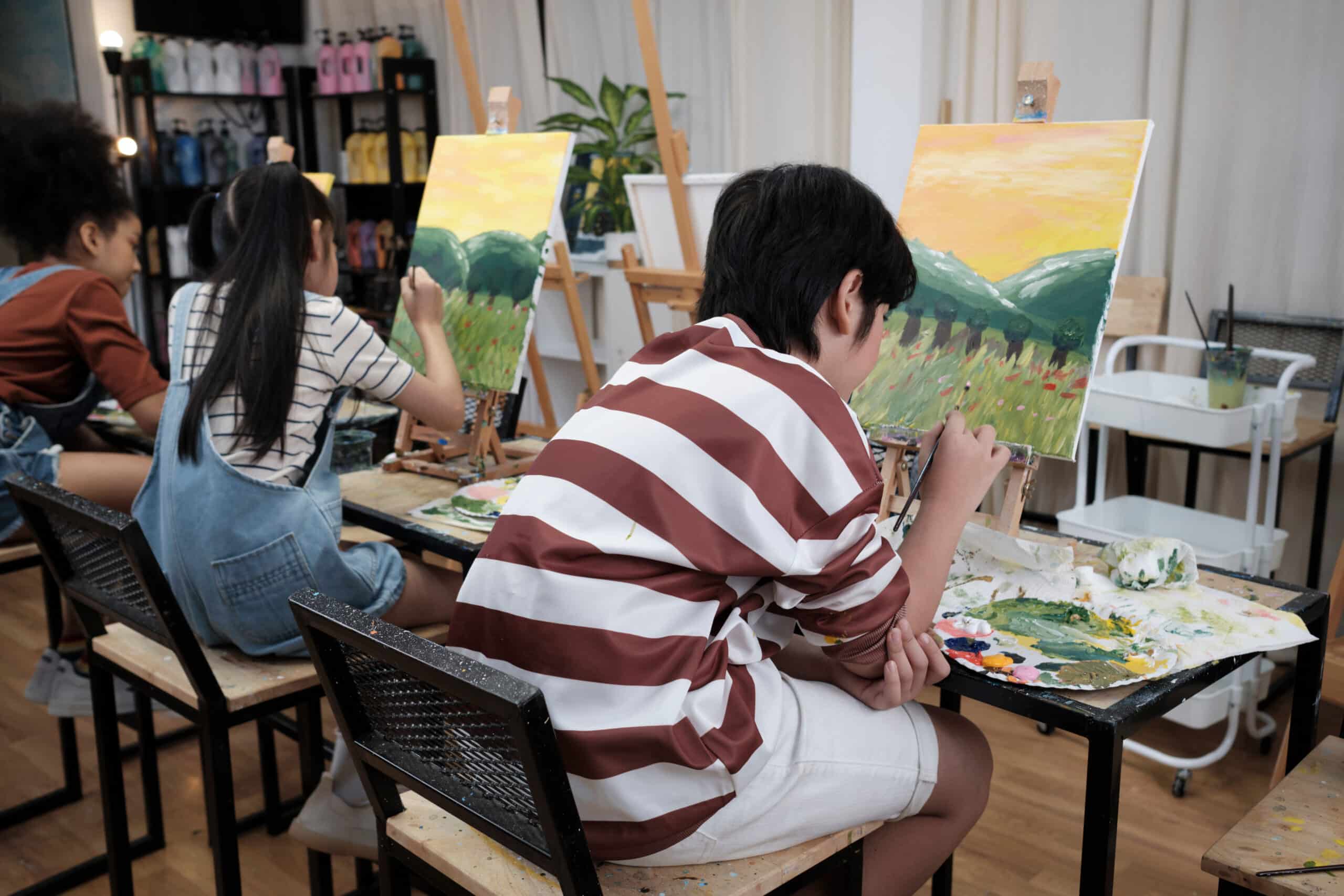 Three children engaged in an art class, painting on easels. One child in a striped shirt sits on a stool, focused on their canvas. Two others, one wearing an overalls and the other in a striped top, work on their paintings, surrounded by art supplies.