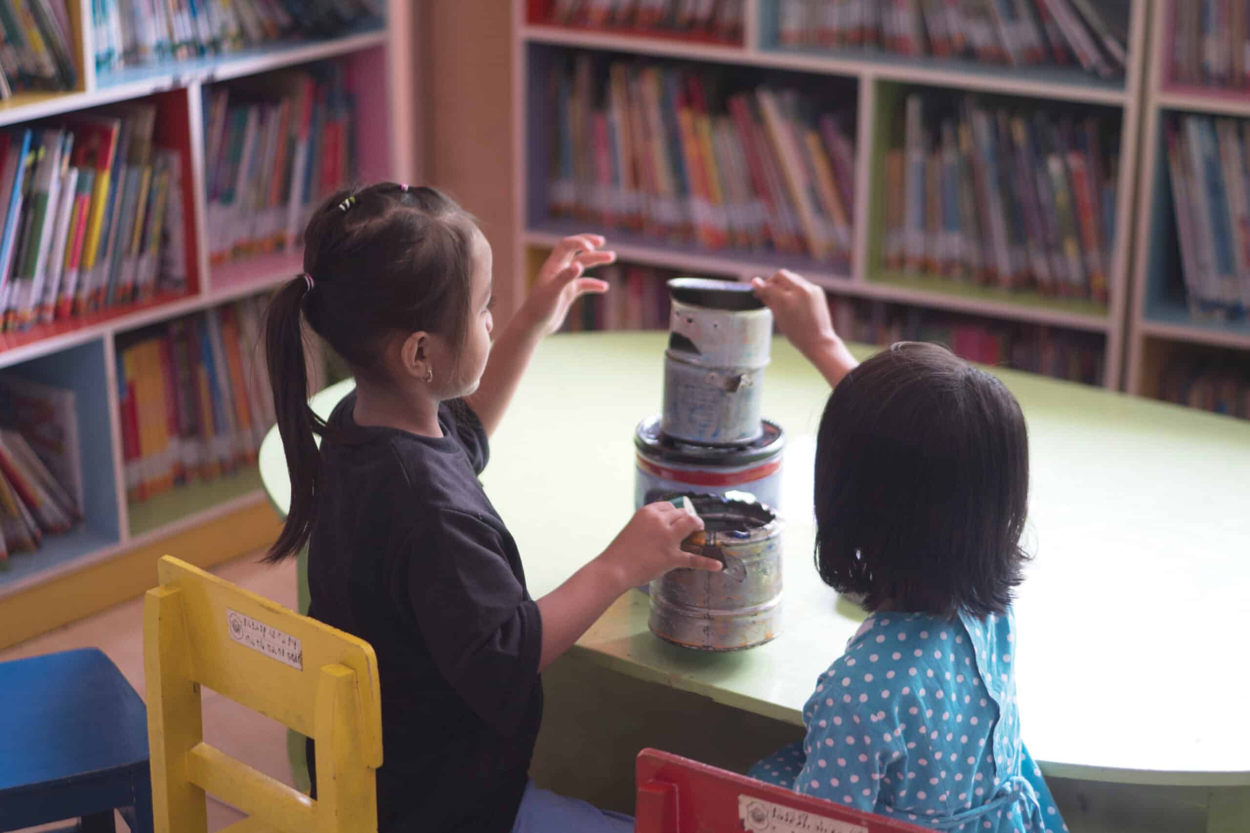 Two children sitting at a round table in a library, actively stacking paint cans. One child wears a black shirt, while the other is in a blue polka dot blouse. Bookshelves filled with books are visible in the background.