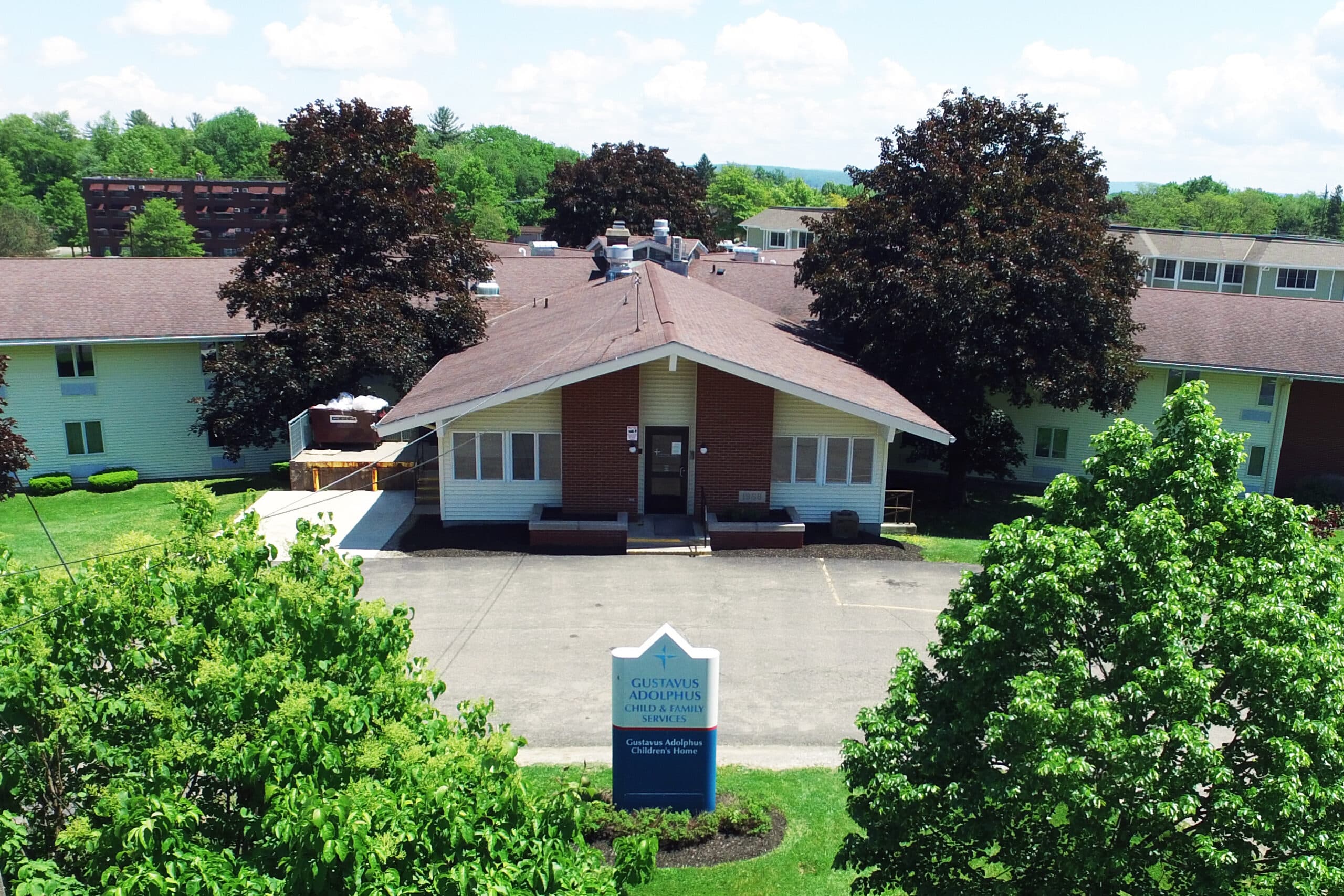 Aerial view of a two-story building with a pitched roof, surrounded by green trees and grass. In front of the building is a sign that reads 'Sapphire Senior Living.' The sky is clear and blue with a few clouds.
