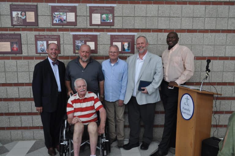 Six men are gathered in front of a brick wall displaying plaques with images and text. One man in a wheelchair, wearing a red and white striped shirt, is seated in the front. The others stand behind him, wearing casual shirts and jackets, smiling at the camera.