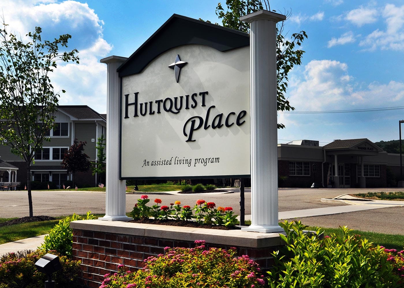 A sign reading "Hultquist Place" with the subtitle "An assisted living program" in a well-maintained outdoor area. The sign is mounted on white pillars, surrounded by colorful flowers and greenery, under a partly cloudy sky.