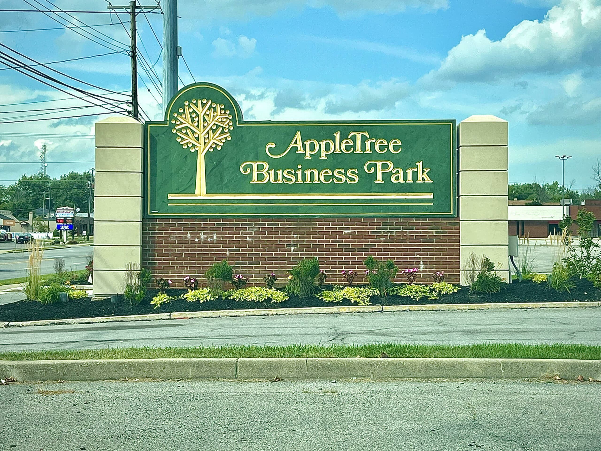 A sign for AppleTree Business Park is displayed prominently, featuring a green background with golden lettering. The sign includes a tree logo and is surrounded by landscaped plants and flowers. Power lines and a cloudy sky are visible in the background.
