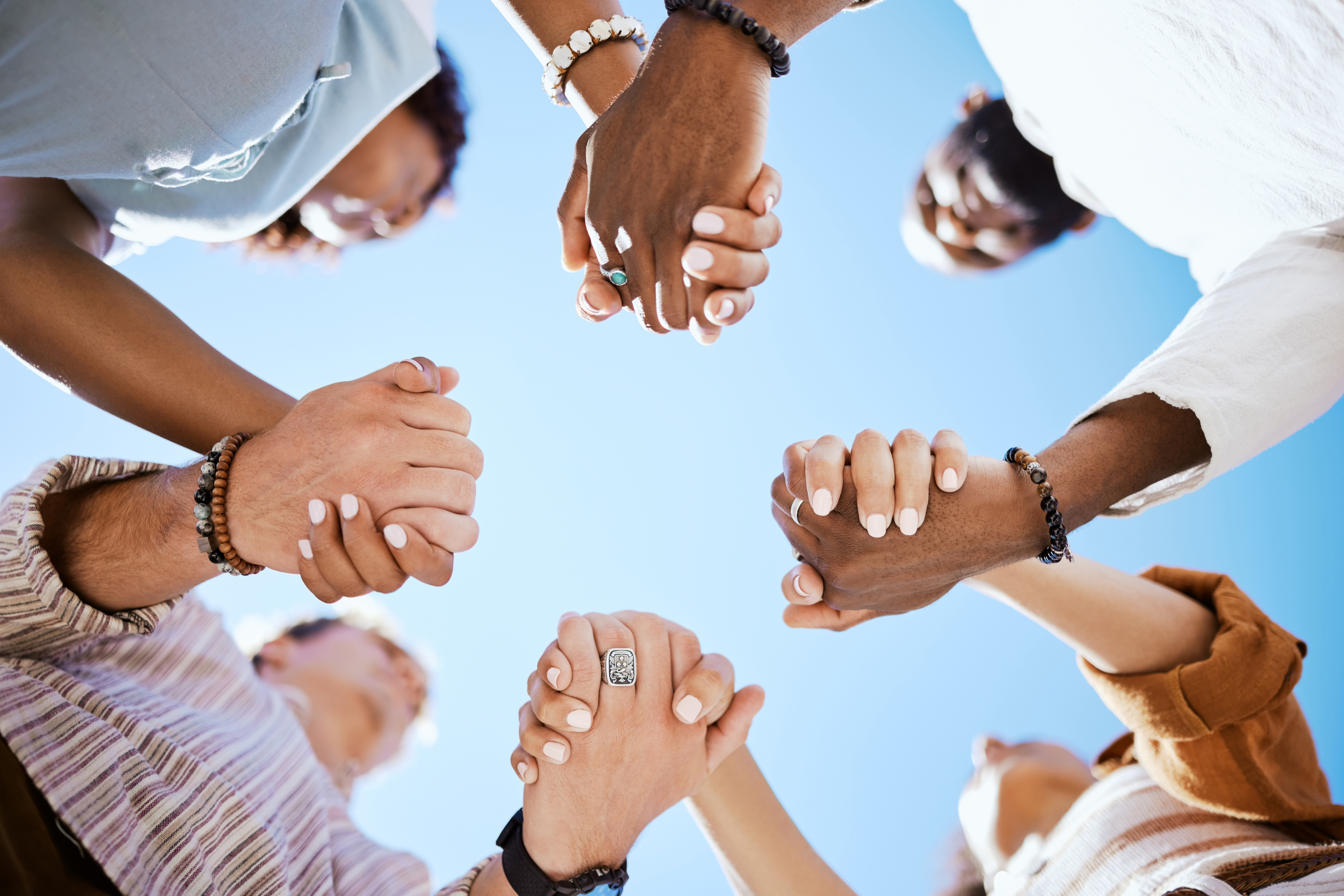 A group of diverse individuals holding hands in a circle against a clear blue sky. Various arm and hand positions are visible, showcasing different skin tones and jewelry, emphasizing unity and connection.