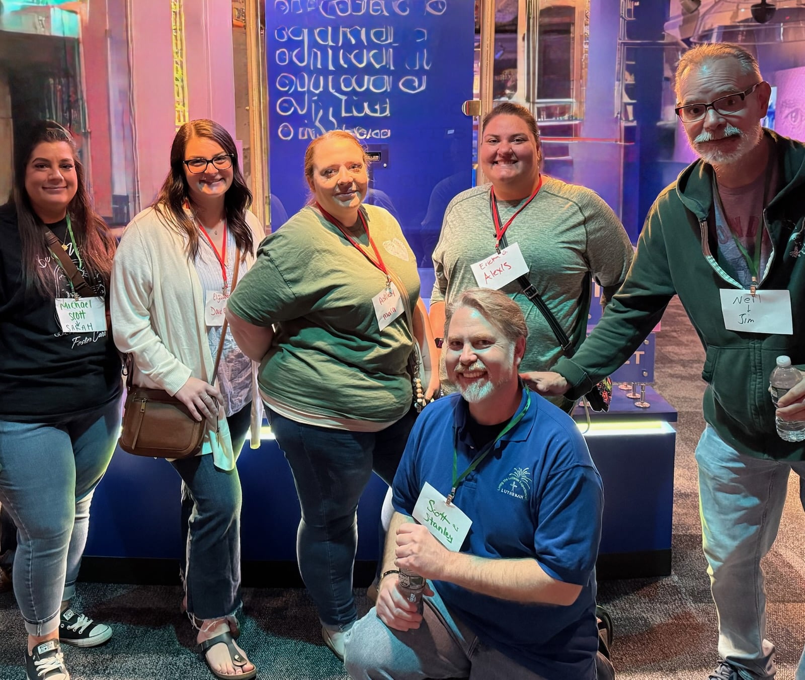 A group of six people posing for a photo in a well-lit exhibition space. They are smiling and wearing name tags. The background features colorful displays and illuminated signage.