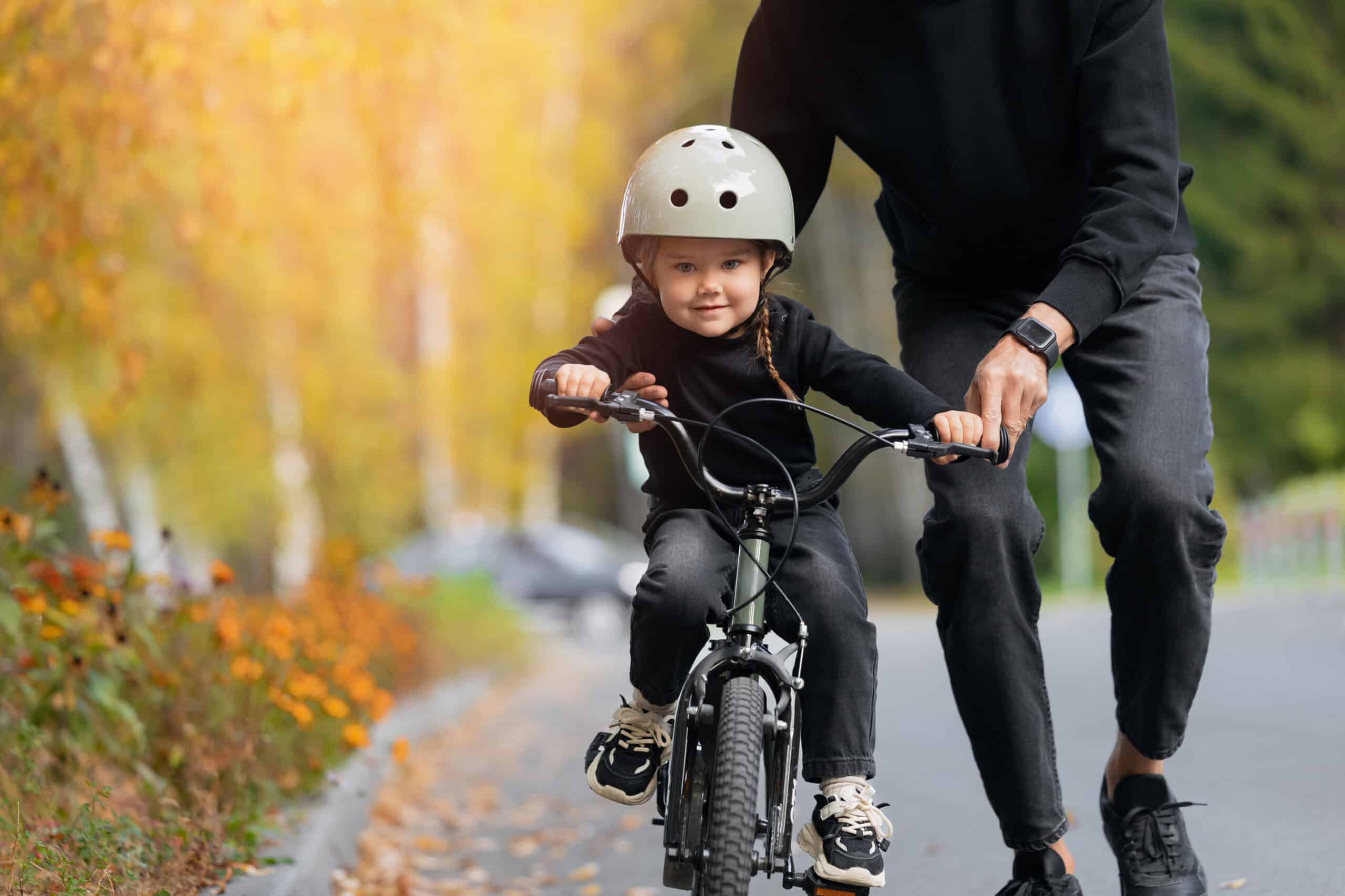A child wearing a helmet rides a small bicycle with assistance from an adult. Both are dressed in black, and the background features trees and autumn foliage. The scene is set on a paved path, suggesting an outdoor activity.