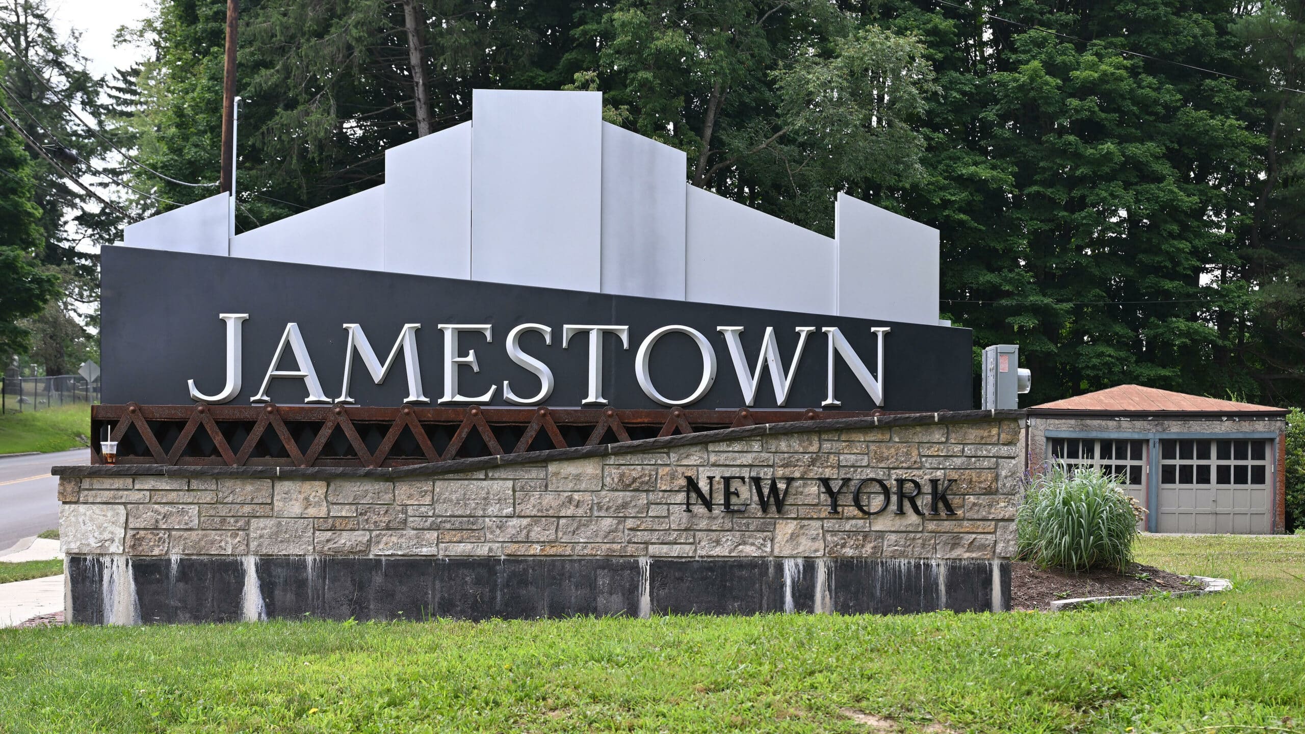 A large sign welcoming visitors to Jamestown, New York, featuring a modern design with the word 'JAMESTOWN' prominently displayed in white against a dark background, and 'NEW YORK' in smaller lettering below, surrounded by greenery.
